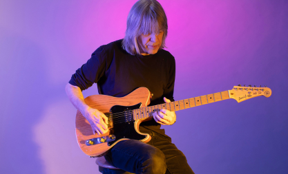 A photo of Mike Stern playing a guitar on a purple-lit background.